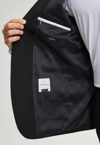 Innenseite eines schwarzen Anzugsakts mit einem glatten, gemusterten Futter. Enthält eine weiße Reißverschlusstasche und Etiketten mit der Aufschrift "Reine Wolle" und dem Markennamen.