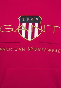 Sudadera roja con un logo bordado en oro "GANT", un escudo con franjas verticales y el texto "1949 AMERICAN SPORTSWEAR".