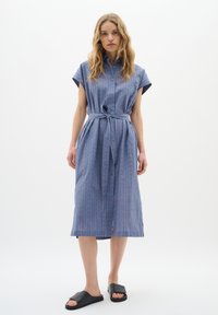 Rochie butonată, cu mânecă scurtă, de culoare albastru deschis, cu curea, având un model texturat și o lungime până la genunchi. Asortată cu sandale negre cu talpă plată.