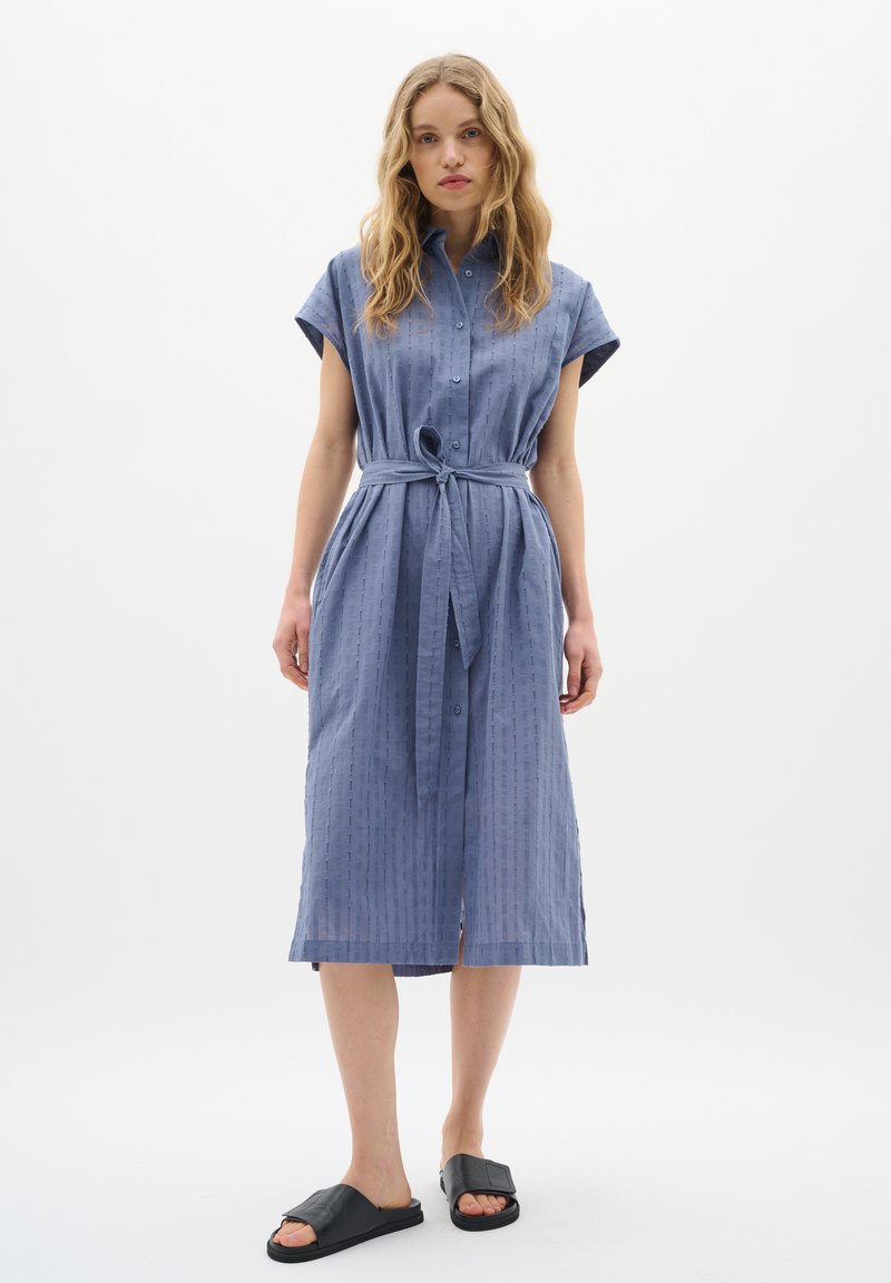 Rochie butonată, cu mânecă scurtă, de culoare albastru deschis, cu curea, având un model texturat și o lungime până la genunchi. Asortată cu sandale negre cu talpă plată.
