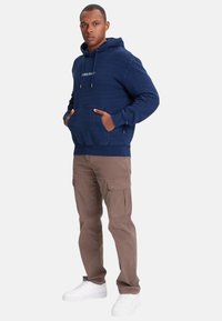 Felpa con cappuccio blu navy con coulisse attaccata, tasca frontale e logo ricamato; abbinata a pantaloni cargo marrone chiaro e sneakers bianche.