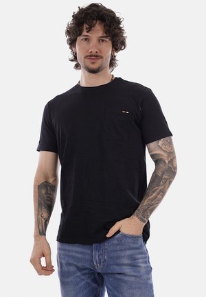 Uomo con capelli ricci che indossa una maglietta nera e jeans blu, con tatuaggi dettagliati su entrambi gli avambracci, una mano in tasca, su sfondo bianco.