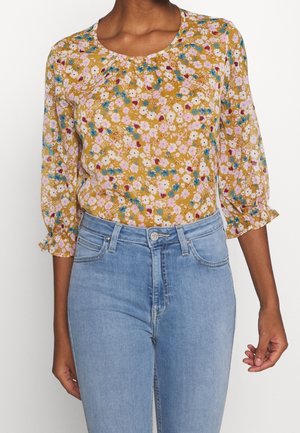 Femme portant un chemisier floral jaune moutarde à manches 3/4 froncées, associé à un jean high-waisted bleu clair.
