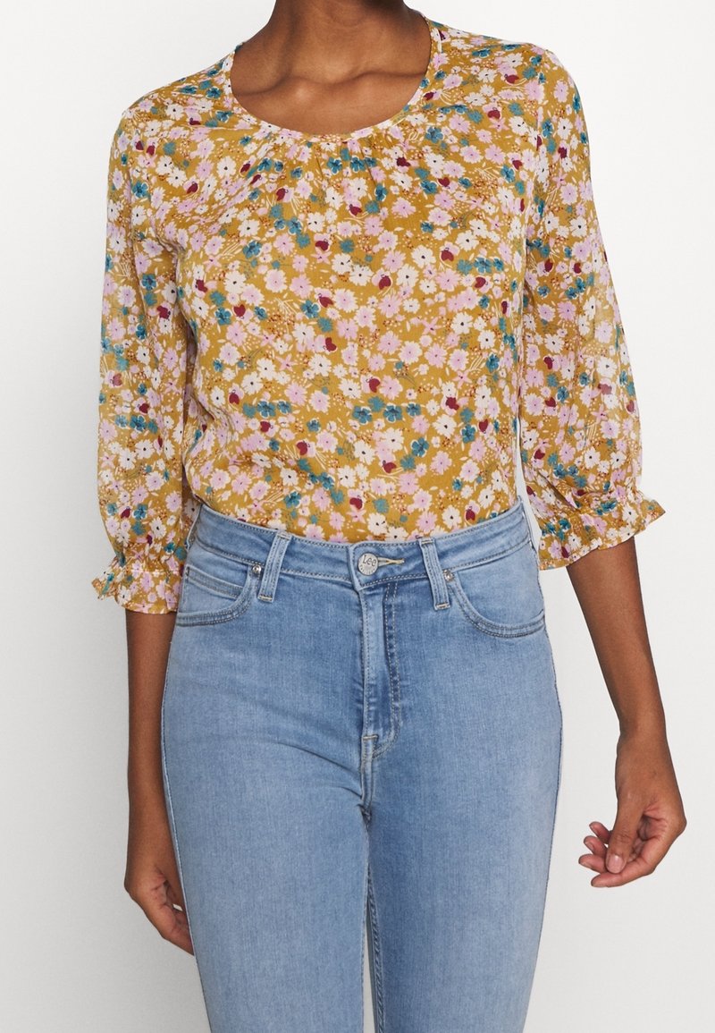 Frau trägt eine senfgelbe Bluse mit Blumenmuster und gerafften 3/4-Ärmeln, kombiniert mit hochgeschnittenen hellblauen Jeans.