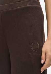 Benešane smeševe hlače v rjavi barvi, ki imajo rebrast vzorec in embosiran logotip z lovorjevimi detajli na strani blizu bokov.