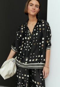 Schwarze Satinbluse mit großen beigen und goldenen Punkten, kurzen Ärmeln und einem Reverskragen. Kombiniert mit passenden Hosen und einer weißen Tasche.