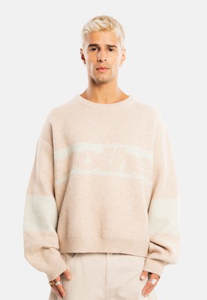 Junge Person mit kurzen blonden Haaren trägt einen beigen, übergroßen Pullover und helle Hosen, steht vor einem schlichten hellen Hintergrund.