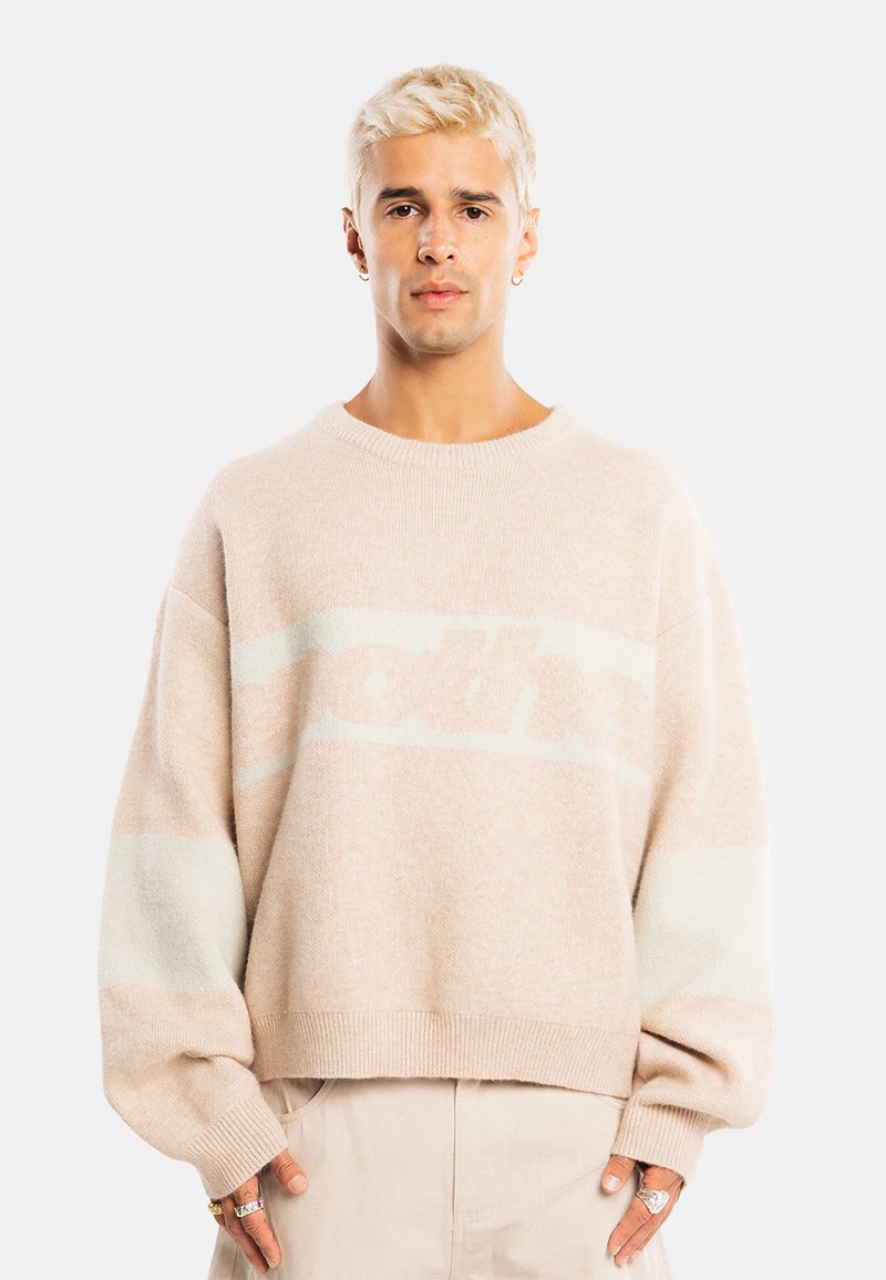 Jeune personne aux cheveux blonds courts porte un pull beige oversize et un pantalon clair, debout devant un fond clair uni.