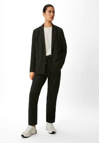Schwarzer, taillierter Blazer mit einem einzigen Knopf, kombiniert mit passenden schwarzen Hosen. Das Model trägt ein weißes Oberteil und hellfarbige Sneakers.