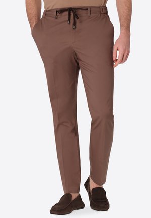 Uomo con pantaloni marroni slim fit con coulisse nera e mocassini marrone scuro, in piedi davanti a uno sfondo semplice.