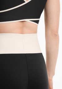 Crni sportski top s bež rubom i odgovarajućim hlačama visokog struka. Ima glatku tkaninu i suptilan logotip na pojasu.