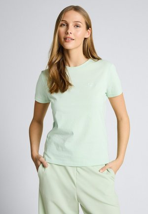 Młoda kobieta ubrana w jasnobeżowy t-shirt i spodnie, stojąca z rękami w kieszeniach na jednolitym tle.