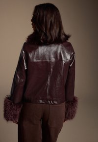 Giacca in pelle bordeaux, polsini rifiniti in pelliccia, design elegante, texture leggermente lucida, lunghezza corta, abbinata a pantaloni marroni.