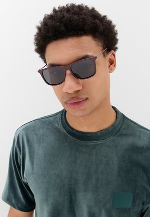Giovane uomo con capelli ricci che indossa occhiali da sole scuri e una maglietta verde a coste a maniche corte su uno sfondo chiaro uniforme.