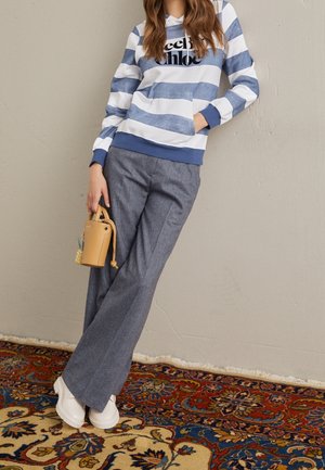 Femme portant un sweat à capuche rayé bleu et blanc, pantalon large gris, chaussures blanches, tenant un petit sac à main beige, debout sur un tapis à motifs.