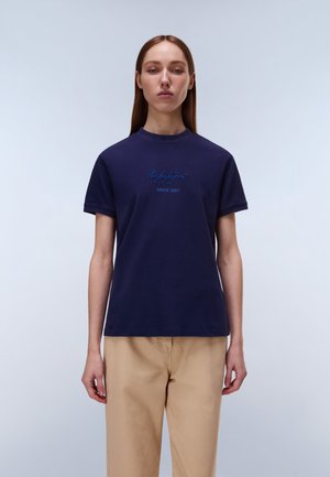 Ung kvinde står oprejst iført en marineblå t-shirt med teksten "Napapijri Since 1987" og beige bukser mod en ensfarvet lys baggrund.