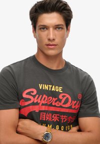 Grå bomulds t-shirt med rød og gul "SuperDry" grafisk tekst. Modellen bærer et guld ur med en marineblå urskive, arme krydset foran.