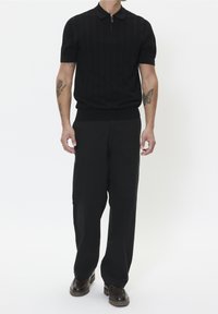 Schwarzes Poloshirt mit kurzen Ärmeln, Kragen und Reißverschluss-Detail, kombiniert mit locker sitzender schwarzer Hose und dunklen Schnürschuhen.