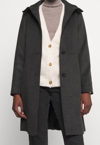 Czarny szary wełniany płaszcz z kapturem, dwoma przednimi kieszeniami oraz zapięciem na guziki. Nałożony na jasny kremowy kardigan z widocznymi złotymi guzikami.