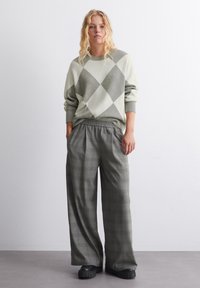 Übergroßer hellgrüner Pullover mit einem Rautenmuster, kombiniert mit grauen weiten Karohosen und schwarzen Plateau-Schuhen.