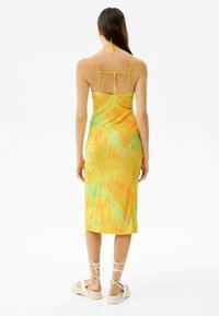 Bezramenné midi šaty v žluté a zelené barvě s motivem palmového listí, nastavitelný halter krk a otevřený detail na zádech, doplněné sandály na tenkých páscích.