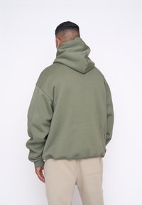 Felpa oversize verde oliva realizzata in tessuto morbido, con un ampio cappuccio e polsini a coste. Abbinata a pantaloni beige dal fit rilassato.