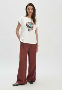 Weißes T-Shirt mit kurzen Ärmeln und buntem botanischen Druck, kombiniert mit weit geschnittenen, rostfarbenen Hosen. Das Model trägt schwarze Ballerinas. Einfacher Hintergrund.