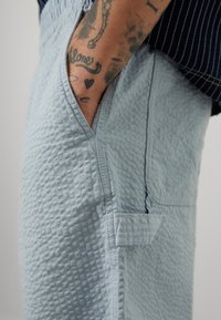 Pantaloni azzurro chiaro con una texture a nido d'ape, dotati di tasche laterali e una piccola etichetta di tessuto rettangolare vicino all'orlo.