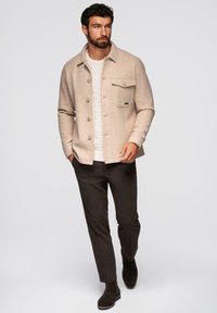 Giacca camicia in lana beige con colletto, tasca frontale, pantaloni scuri e stivali in suede marrone indossati sopra un maglione bianco a trecce.