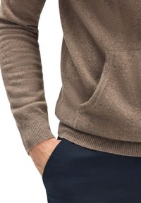 Brun, struktureret sweater med lange ærmer og en frontlomme, parret med mørkeblå bukser. Ribbekanter og detaljer ved kanten er synlige.