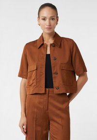 Veste courte marron à manches courtes, avec deux poches avant et une fermeture à bouton. Tissu lisse et léger. Associée à un pantalon assorti.