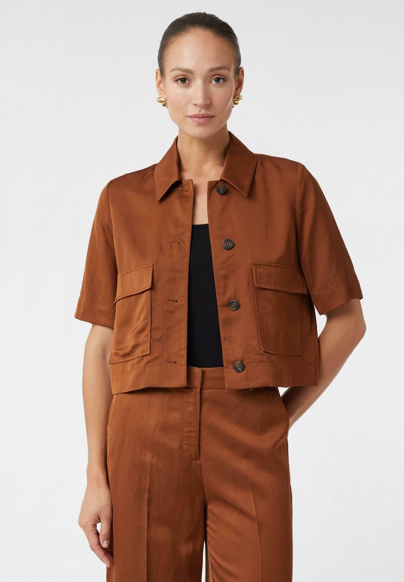 Veste courte marron à manches courtes, avec deux poches avant et une fermeture à bouton. Tissu lisse et léger. Associée à un pantalon assorti.