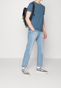 Persona che indossa una maglietta blu, jeans azzurri chiari, sneakers grigio e bianche, con uno zaino nero a spalla, in piedi su uno sfondo neutro.