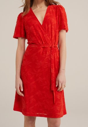 Femme portant une robe portefeuille rouge mi-longue à manches courtes avec un motif floral en relief, attachée à la taille par une ceinture.