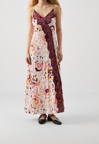 Maxi sukienka na ramiączkach typu spaghetti, z kwiatowym wzorem w odcieniach pomarańczowego i fioletowego na białym tle oraz bordowymi koronkowymi akcentami.