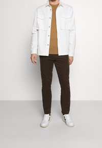 Camicia bianca con bottoni sopra un maglione marrone, abbinata a pantaloni in velluto a coste marroni e sneakers bianche con dettagli neri. Outfit semplice e informale.