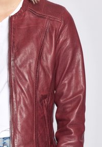 Giacca in pelle rossa con finitura lucida, dotata di colletto a coste, chiusura con zip e tasche laterali. La vista ravvicinata mette in risalto la texture e i dettagli.