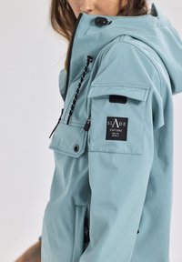 Giacca impermeabile azzurro chiaro con zip, cappuccio, molteplici tasche e branding nero sulla manica. Presenta cordini regolabili e tessuto testurizzato.