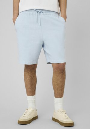 Homme portant un short bleu clair à cordon, des chaussettes blanches et des baskets blanc cassé, debout avec les mains dans les poches devant un fond gris.