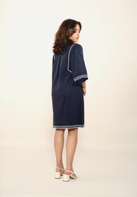 Navyblaues Kleid mit kurzen Puffärmeln, das weiße Stickereien am Saum und an den Ärmeln aufweist. Die Stoffstruktur wirkt weich und leicht.