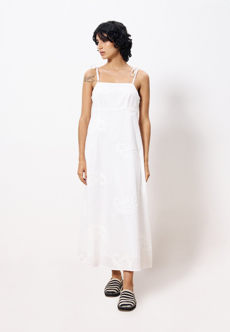 Weißes Maxikleid mit floraler Stickerei, dünnen Trägern und tailliertem Oberteil. Kombiniert mit schwarz-weißen gestreiften Slippern.