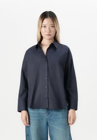 Young woman with straight blonde hair wearing a dark navy button-up shirt and light blue denim jeans, standing against a white background.