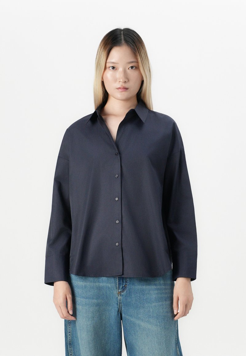 Young woman with straight blonde hair wearing a dark navy button-up shirt and light blue denim jeans, standing against a white background.
