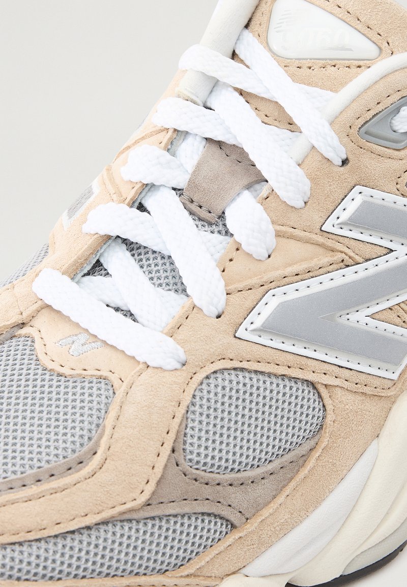 Beige und graue Sportschuhe aus Mesh- und Wildledermaterialien, mit weißen Schnürsenkeln, silbernen Akzenten und einer runden, gepolsterten Sohle.