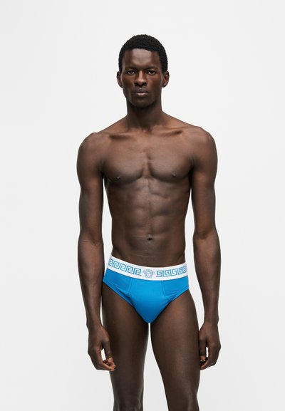 Homme debout face à l’avant portant un slip bleu avec une ceinture blanche à motifs, sur un fond uni clair.