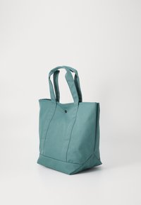 Teal tote taske med to solide håndtag, rektangulær form, lavet af slidstærkt stof, har en sort dekorativ knap på forsiden.