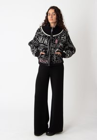 Zwarte ritstrui met een oversized kraag, voorzien van geometrische witte en roze patronen. Gecombineerd met zwarte wijde broek.