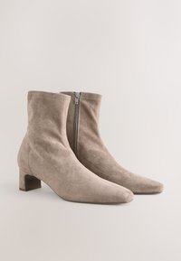 Stivaletti alla caviglia in camoscio beige con punta appuntita, chiusura laterale con zip e tacco a blocco modesto. Texture liscia e design semplice.