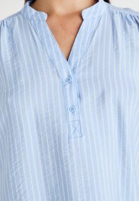 Camisa de color azul claro con cuello y rayas verticales blancas, que presenta un cierre de botones y una textura sutil. Los botones coinciden con el color de la tela.