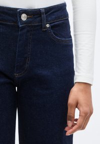 Σκούρο μπλε τζιν με ψηλή μέση, με ραφές αντίθετου χρώματος, κούμπωμα με κουμπί μπροστά και πλαϊνές τσέπες. Το χέρι ακουμπά δίπλα στο τζιν.